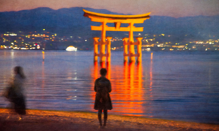 Two tourists, one walking and one standing, in front of illuminated O-torii with lights in Hiroshima in the background